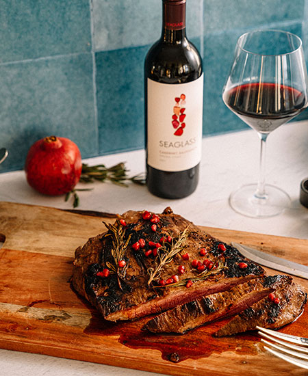 Sliced flank steak lies on a wooden chopping board next to a whole pomegranate, a sprig of rosemary, a wine glass, and a bottle of SEAGLASS Cabernet Sauvignon